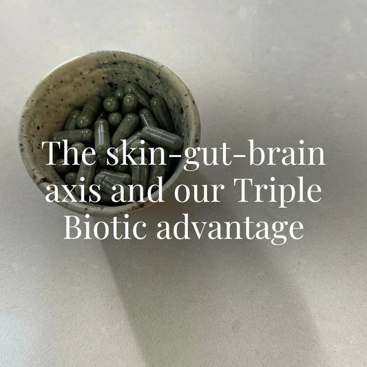 A ceramic bowl filled with greenish-black supplement capsules sits on a clean cream-colored surface. The image is simple, functional, and natural. The overlaid text reads: “The skin-gut-brain axis and our Triple Biotic advantage.”